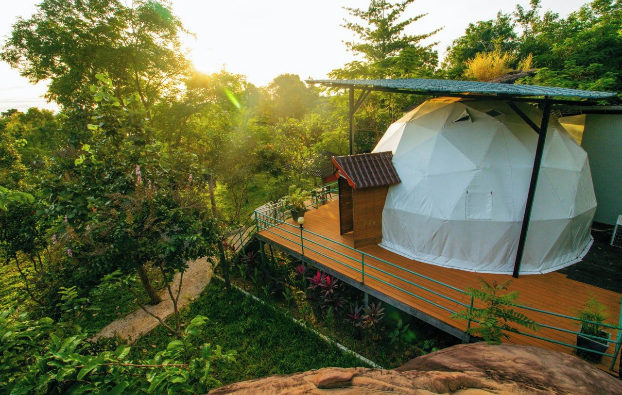 A tented accommodation with a wooden deck and a small building with a sloped roof, surrounded by lush greenery.