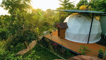 A tented accommodation with a wooden deck and a small building with a sloped roof, surrounded by lush greenery.