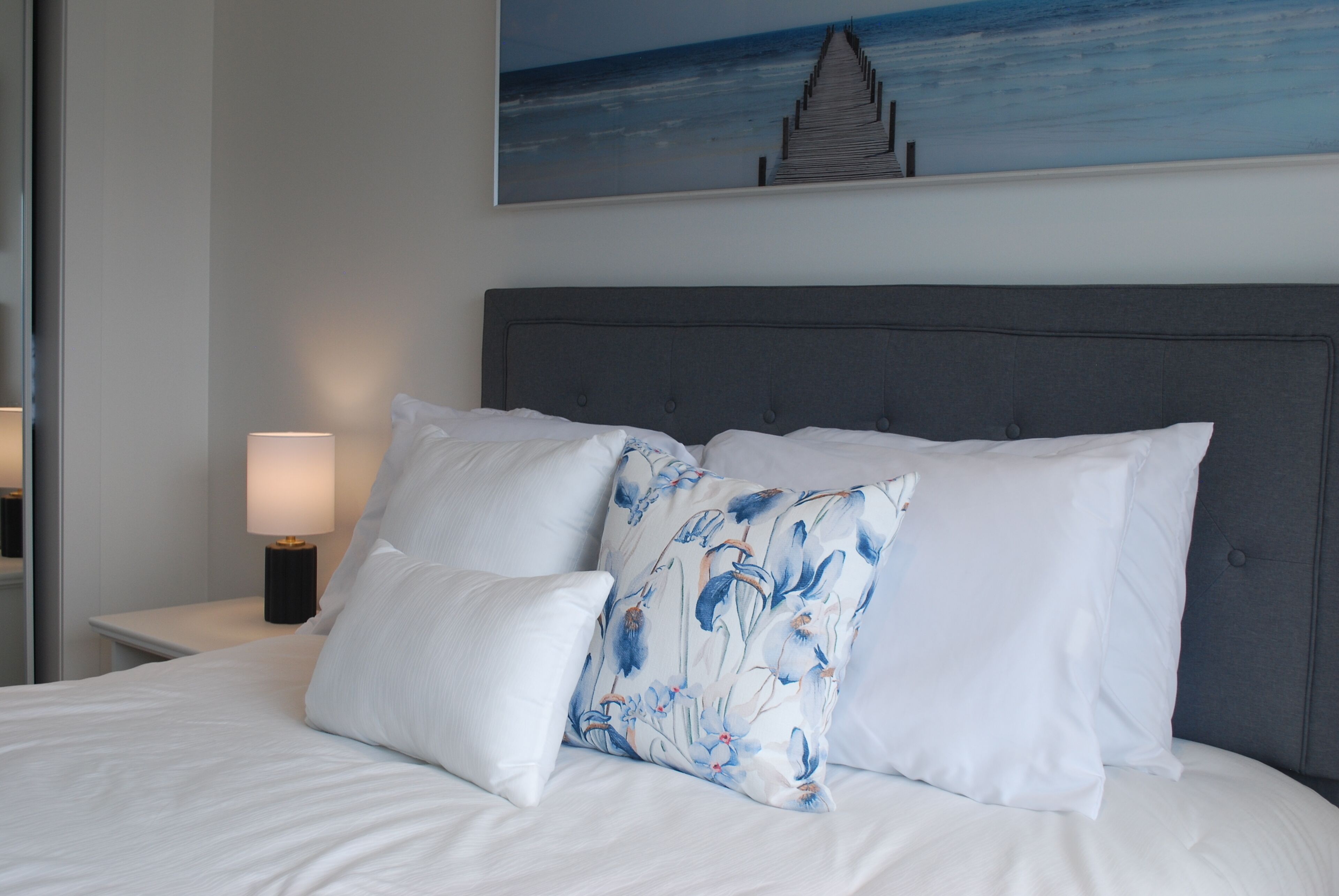 A bed with white bedding and a grey headboard, a nightstand with a lamp, and a painting of a pier on the wall.