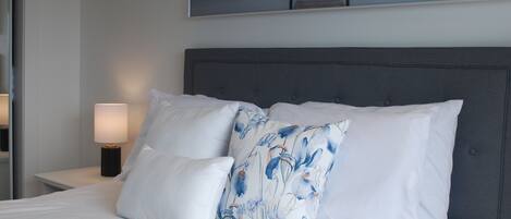 A bed with white bedding and a grey headboard, a nightstand with a lamp, and a painting of a pier on the wall.