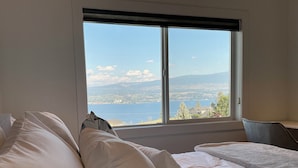 Une chambre avec une grande fenêtre donnant sur un plan d’eau et sur une étendue de terre au loin. On y trouve deux lits avec du linge de lit blanc et une chaise.