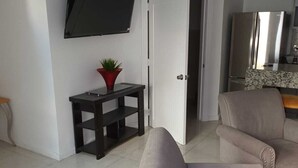 Ett vardagsrum med en platt-TV som är monterad på väggen, ett litet bord med en växt, en grå fåtölj och en köksdel med ett kylskåp.