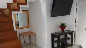 Ett modernt vardagsrum med en trätrappa, en platt-TV som är monterad på väggen, ett litet bord med en växt och en grå soffa.