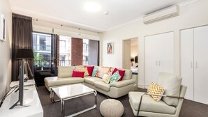 A modern living room with a sectional sofa, a glass coffee table, a round ottoman, and a chair. There is a lamp, a TV, and a wardrobe. - Subiaco Rooftop on Sheen (Subiaco)