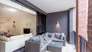A modern living room with a white sofa, grey sectional sofa, and wicker furniture. There is a TV, a small table, and a wall-mounted light fixture. - Subiaco Rooftop on Sheen (Subiaco)