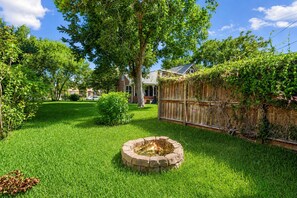 A backyard with a stone fire pit surrounded by grass, a wooden fence, and a house in the background.