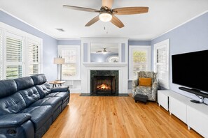A living room with a fireplace, a leather sofa, a patterned armchair, a television, and a ceiling fan. - Game Room | Fenced Yard | Fire Pit | Sleeps 8 (Galveston)
