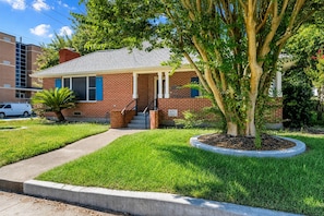 A two-story brick house with a front porch, a tree in the front yard, and a sidewalk leading to the house. - Game Room | Fenced Yard | Fire Pit | Sleeps 8 (Galveston)