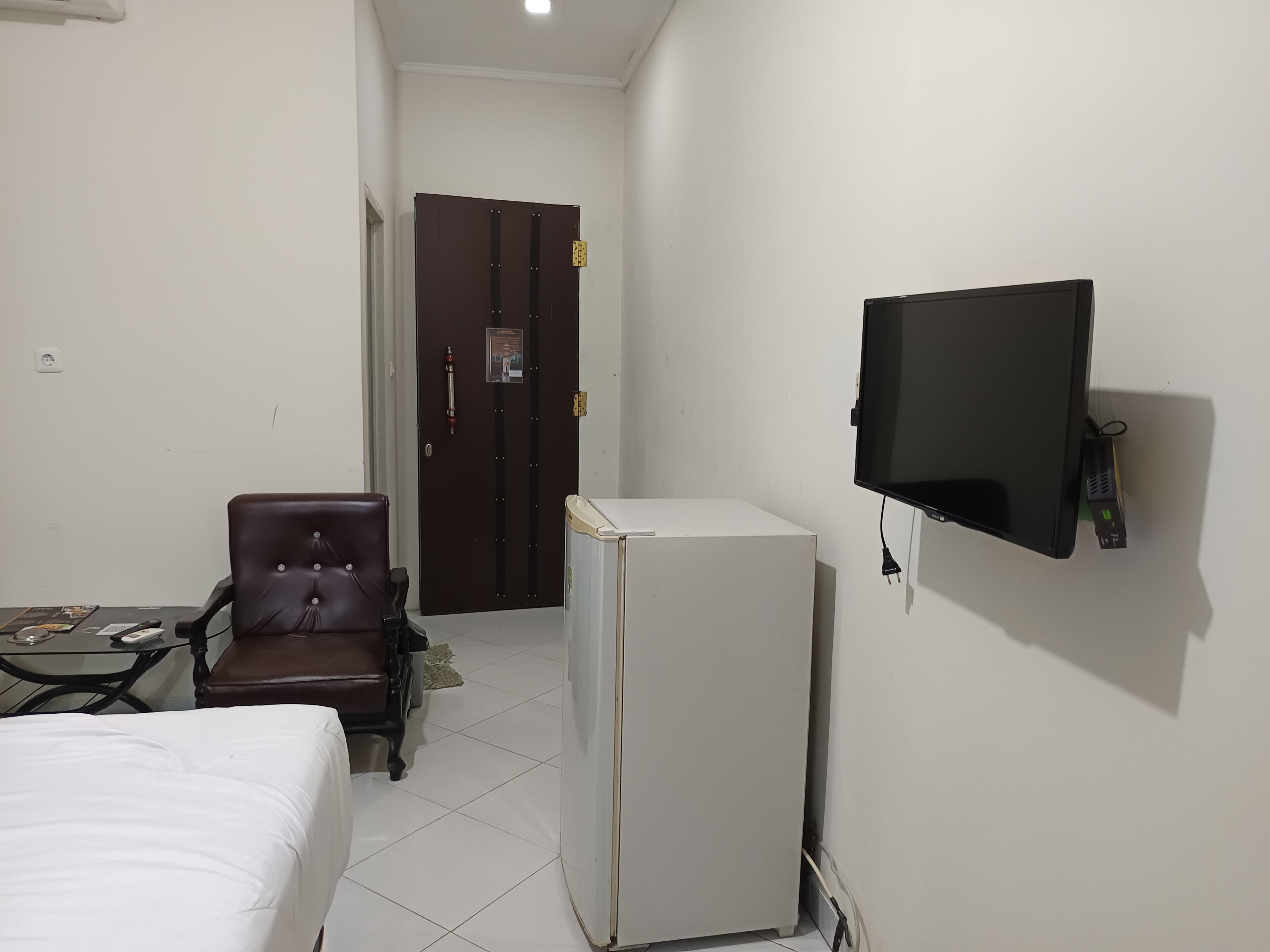 Habitación de hotel con cama, escritorio con silla, refrigerador, televisor montado en la pared y puerta con extintor.