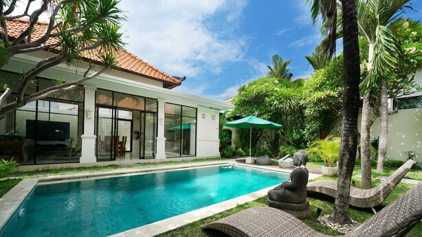 A swimming pool with lounge chairs and a statue, surrounded by greenery and a building with large windows.