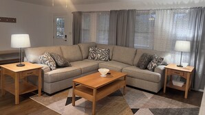 Ein Wohnzimmer mit Ecksofa, zwei Beistelltischen mit Lampen, einem Deckenventilator und einem gemusterten Teppich.