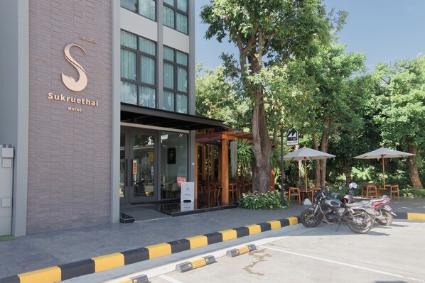 En hotelindgang med et skilt, der viser "Sukruethai Hotel", udendørs siddepladser og en parkeringsplads med motorcykler.