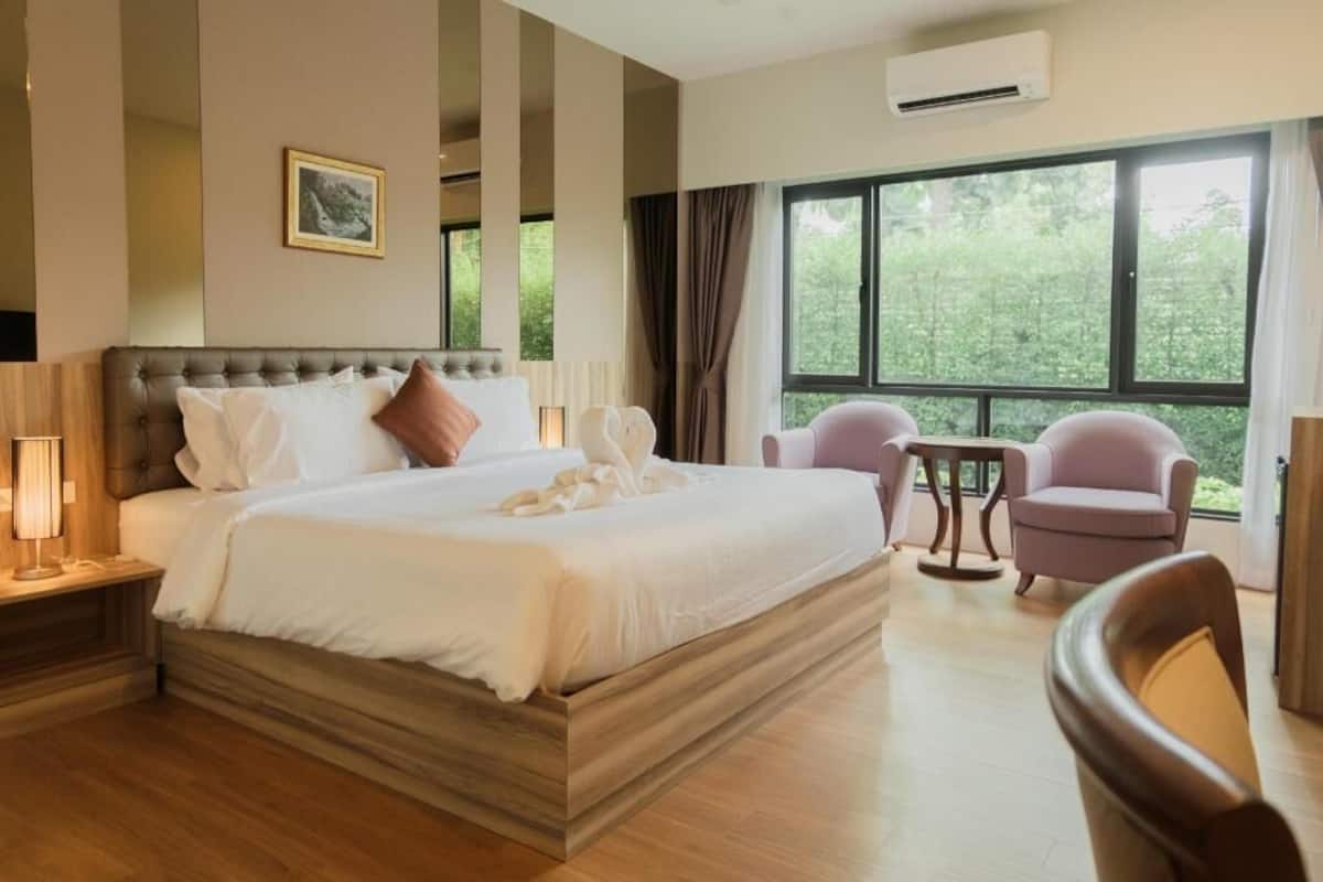 A hotel room with a large bed, two armchairs, a small table, and a window with a view of greenery.