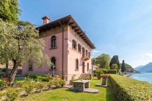 Une villa rose dotée d’un balcon, d’un banc en pierre et d’un jardin agrémenté de fleurs rouges.