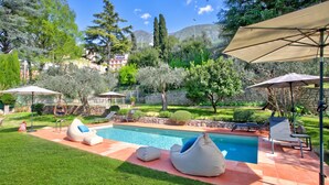 A swimming pool with lounge chairs and umbrellas, surrounded by a well-maintained garden and trees.