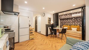 A compact kitchen with a refrigerator, stove, and sink. There is a dining area with a table and chairs, and a sofa with cushions. - Designer-style 2-bedroom apartment & large living room Max 7people Shinjuku 501 (新宿区)