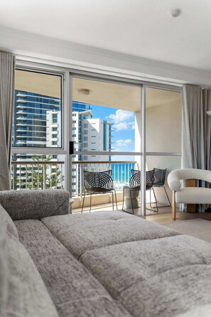 Ein modernes Hotelzimmer mit einer großen Schiebeglastür, die auf einen Balkon führt. Das Zimmer ist mit einem grauen Sofa, einem weißen Sessel und zwei schwarzen Stühlen ausgestattet. Der Balkon bietet Blick auf hohe Gebäude und einen klaren Himmel.