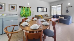 A dining area with a round wooden table set for four, surrounded by wooden chairs. There is a sofa and a window with curtains in the background.