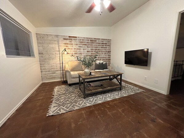 A living room with a brick wall, a sofa, a coffee table, a floor lamp, and a TV mounted on the wall. - Chula Vista Cottage - Fully stocked, clean and central 1 bed, 1 bath in Tucson (Tucson)
