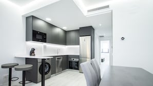 Una cocina moderna con armarios grises, horno empotrado y refrigerador de acero inoxidable. Hay dos taburetes de bar y una mesa de comedor con sillas grises.