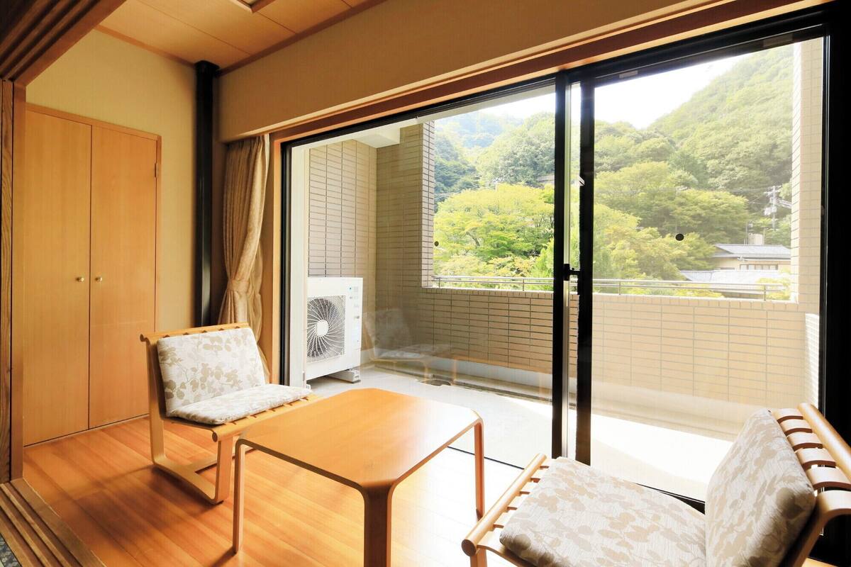 A room with a sliding glass door leading to a balcony with a view of a forested area. There is a wooden table and two chairs with cushions.