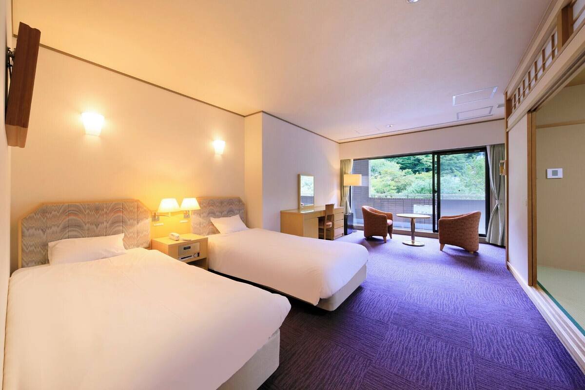 A hotel room with two beds, a desk, a chair, and a view of the outdoors.