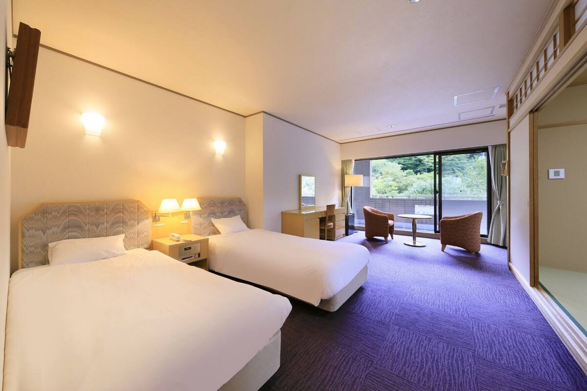 A hotel room with two beds, a desk, a chair, and a view of greenery through the glass door.