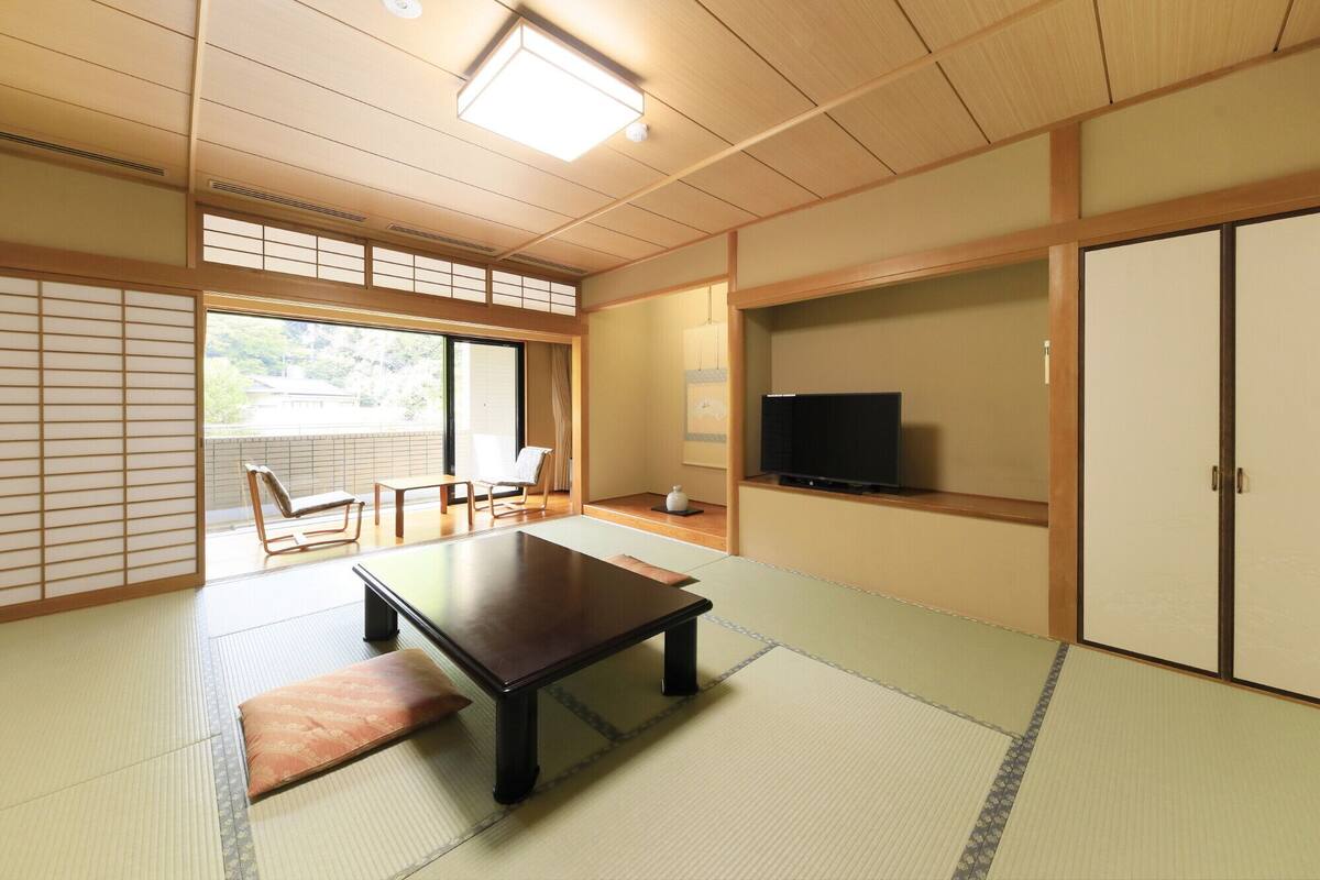 A traditional Japanese room with tatami flooring, a low table, a TV, sliding doors, and a view through a large window.