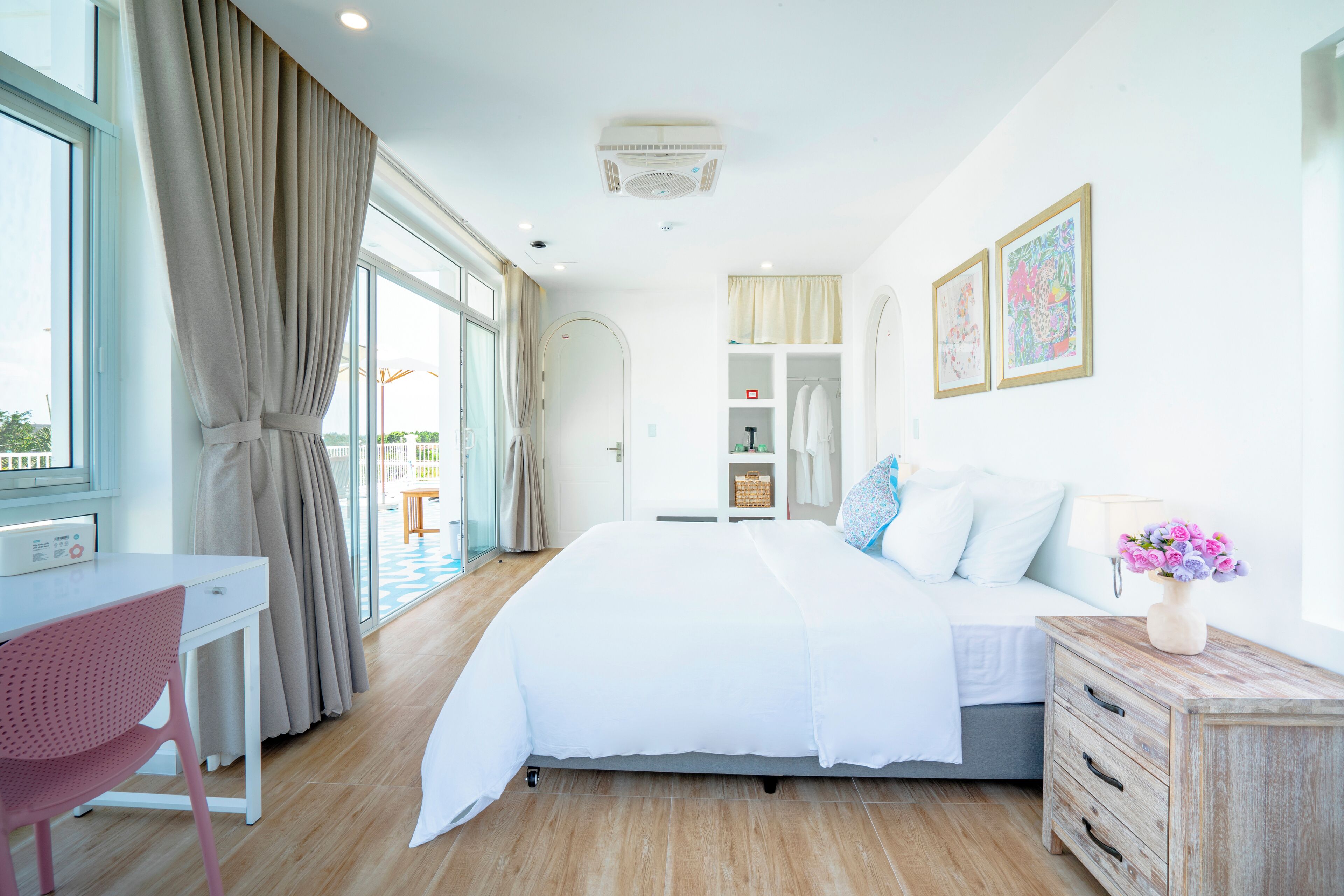 A bedroom with a bed, a desk, a chair, a bedside table with a vase, and a view of the outdoors.