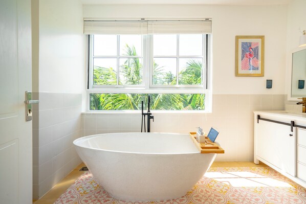A modern bathroom with a freestanding bathtub, a window with a view of greenery, and a small wooden tray with toiletries.