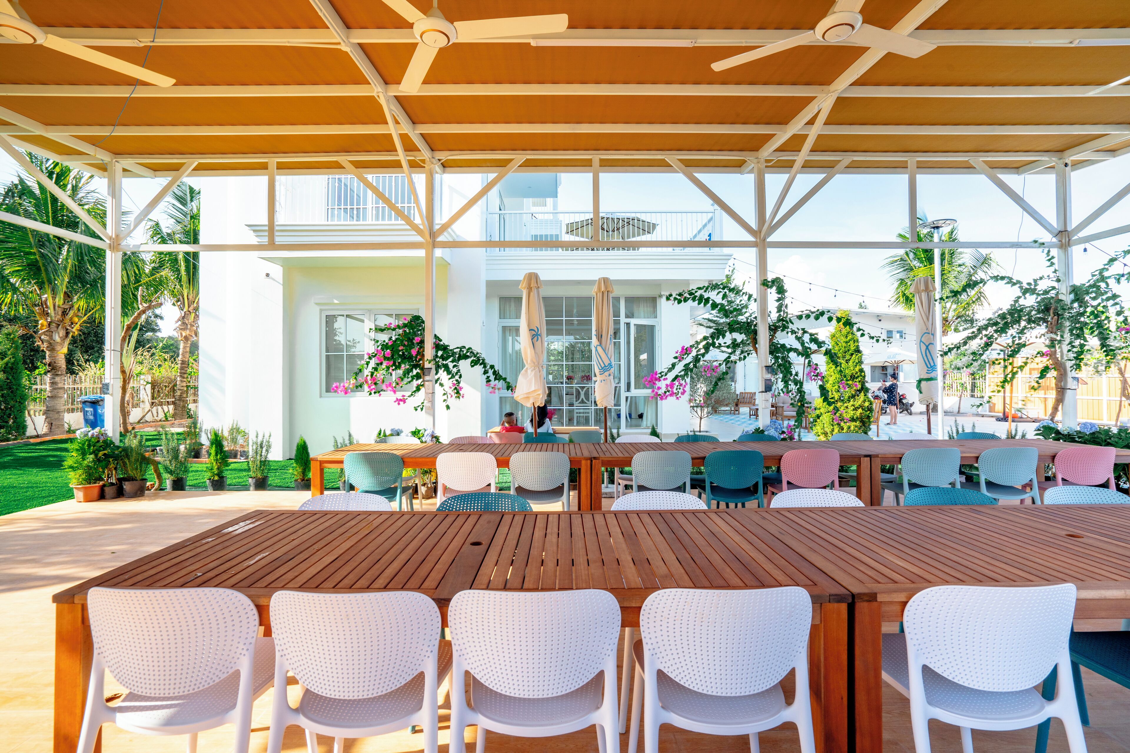 An outdoor patio area with a wooden table and white chairs, surrounded by potted plants and umbrellas.