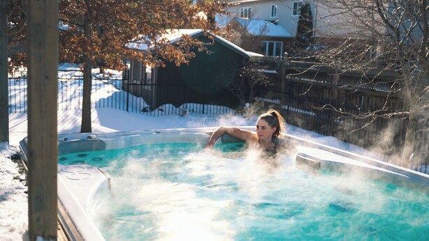 A person in a hot tub with steam rising, surrounded by a snowy landscape and a wooden fence.