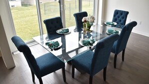 A dining area with a glass table and blue upholstered chairs, a vase with white flowers, and a view of a grassy yard through large windows.