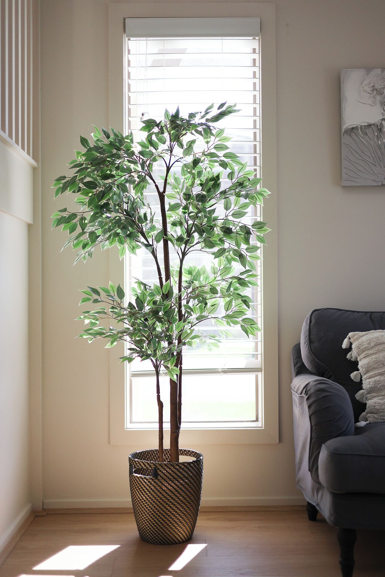 A room with a large window, a dark grey sofa, and a woven basket containing a green artificial tree.