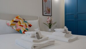 A neatly made bed with white towels, a colorful geometric pillow, a vase with pink flowers, and a framed picture on the wall.