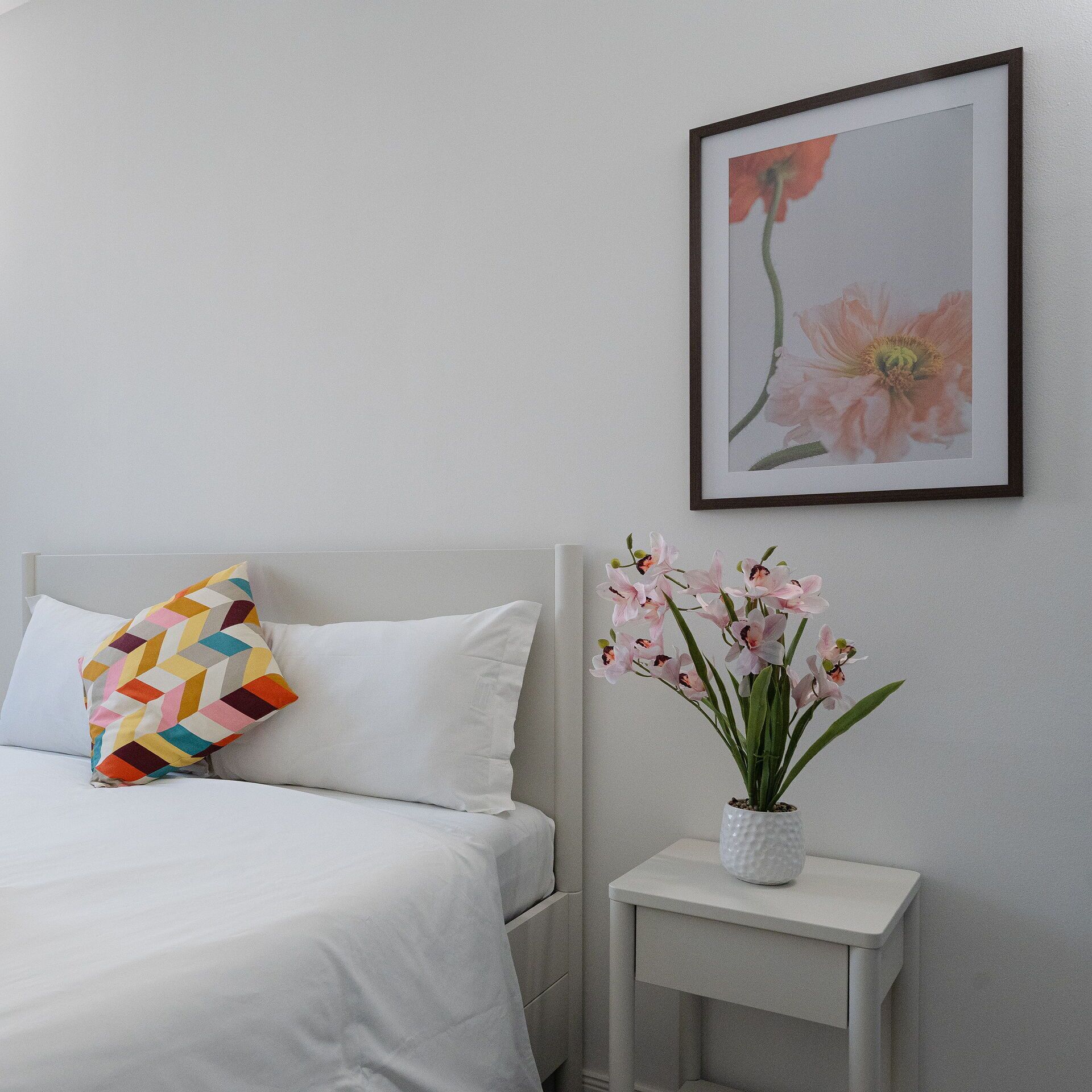 A neatly arranged bedroom with a bed, pillows, a bedside table with a vase of flowers, and a framed picture on the wall.