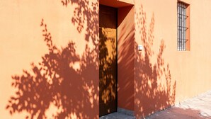 A building corner with a wooden door, a window with bars, and shadows cast by trees.