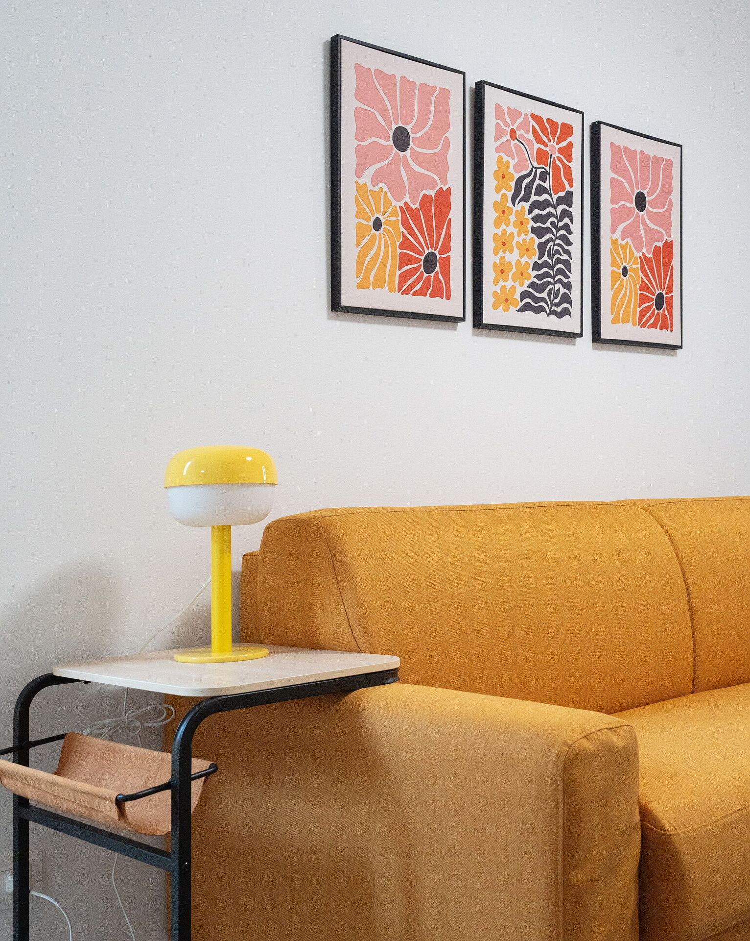 A room with a mustard-colored sofa, a small table with a yellow lamp, and three framed abstract art pieces on the wall.