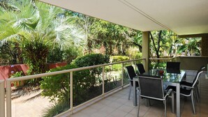 A balcony with a glass table and chairs, surrounded by lush greenery and a view of a garden. - Beachfront Location -Ground Floor. Listen to the waves! (Mooloolaba)