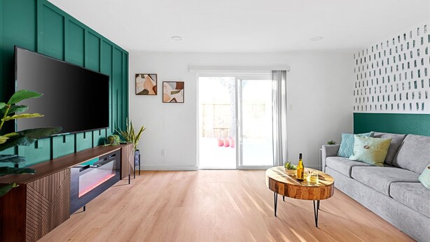 Ein modernes Wohnzimmer mit Holzboden, einem flachbildfernseher an einer türkisblauen Wand, einem grauen Sofa und einem runden Couchtisch mit Flasche und Gläsern.
