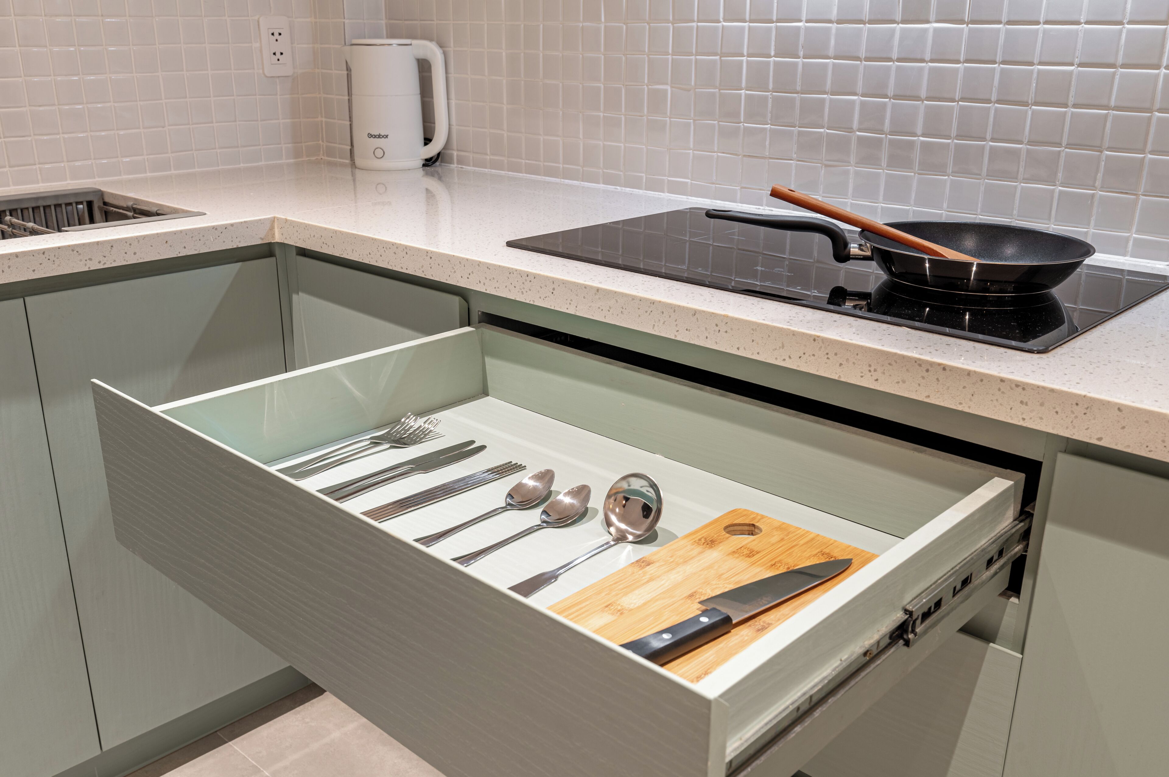 A kitchen with a drawer containing utensils, a cutting board, and a frying pan on a stove.