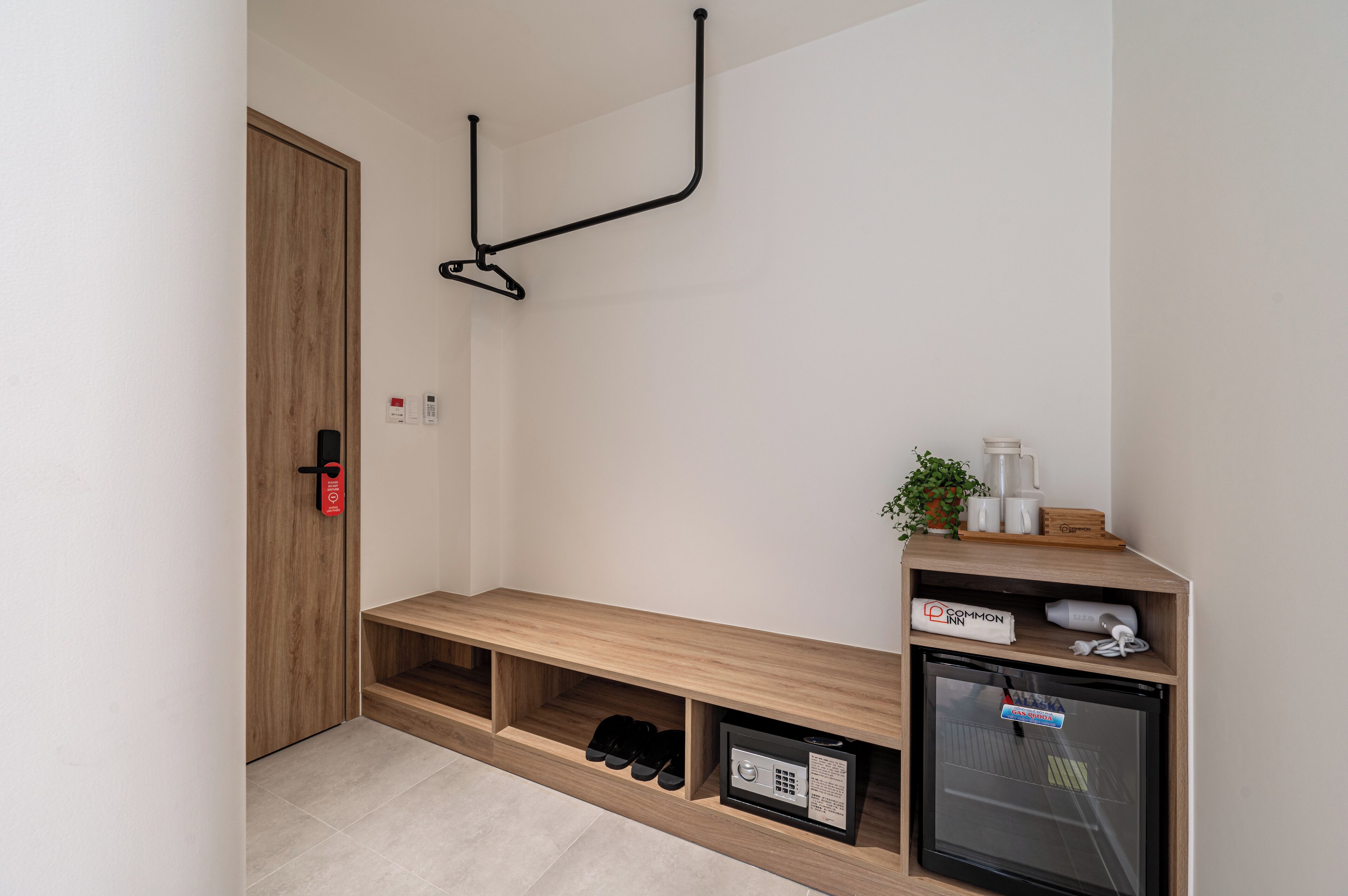 A hotel room with a wooden bench, a small refrigerator, a shoe rack, and a wall-mounted hook.