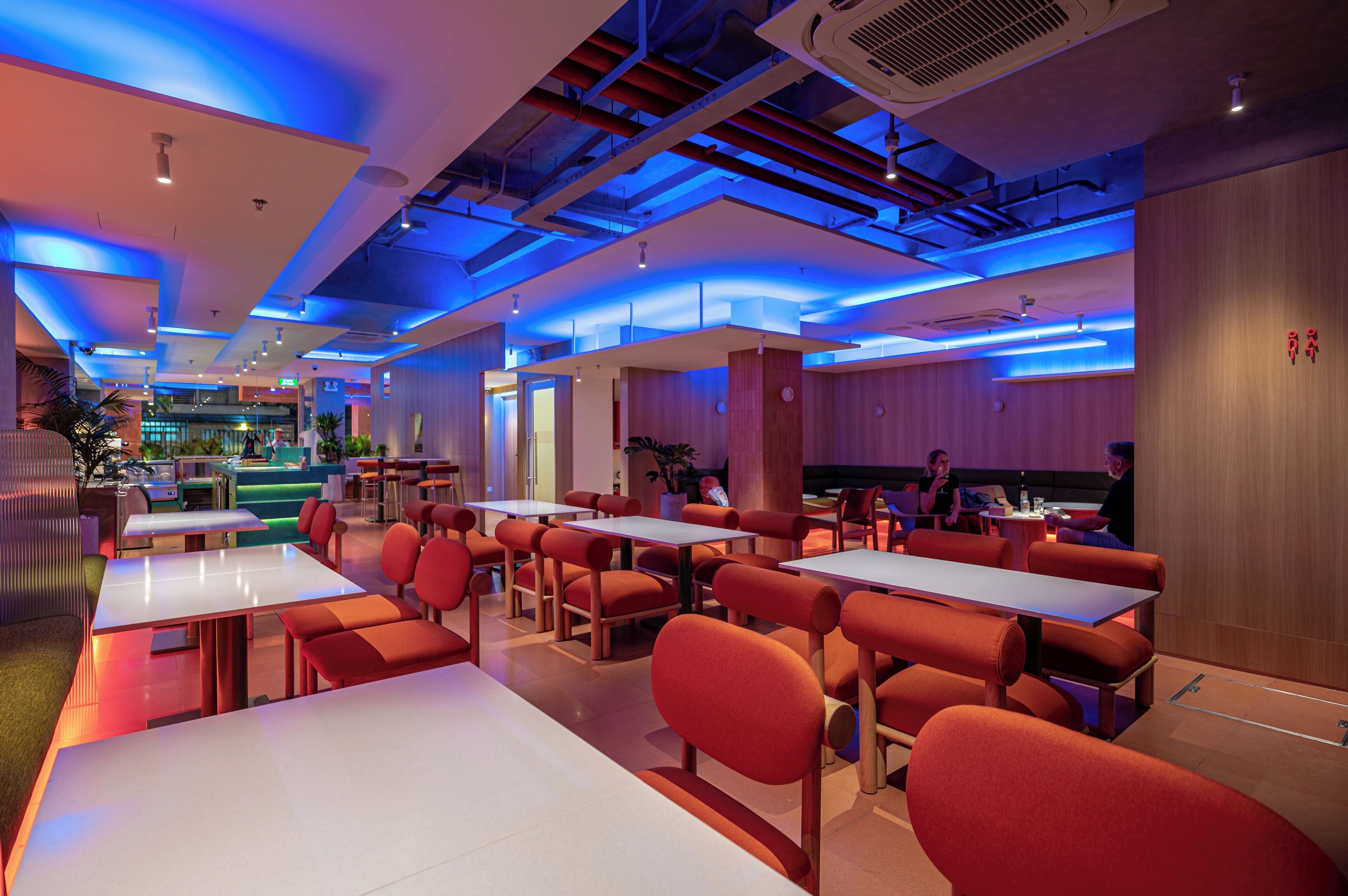 A modern restaurant with white tables and orange chairs, featuring a ceiling with blue lighting and visible ductwork.