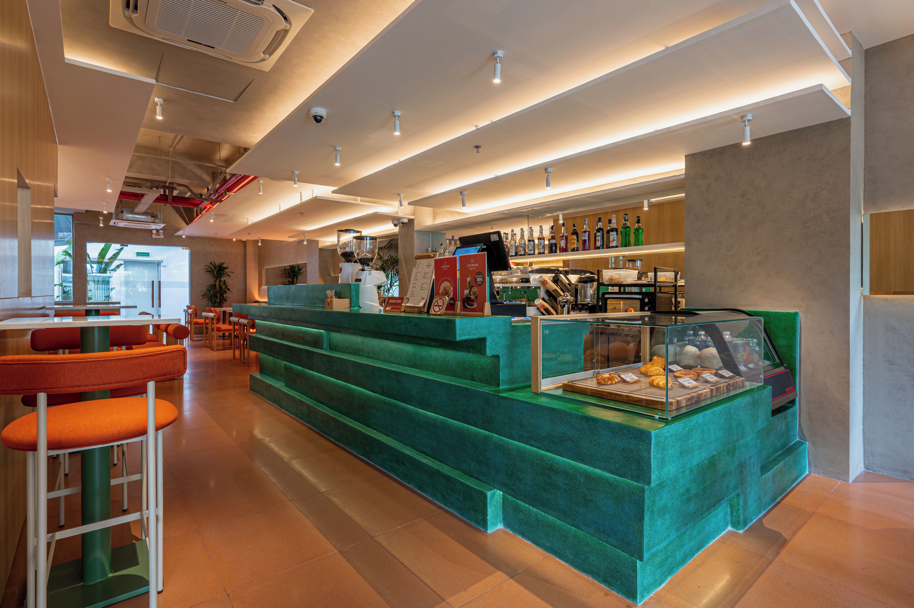 Zona de bar moderna con mostradores verdes, taburetes y una vitrina con pasteles.