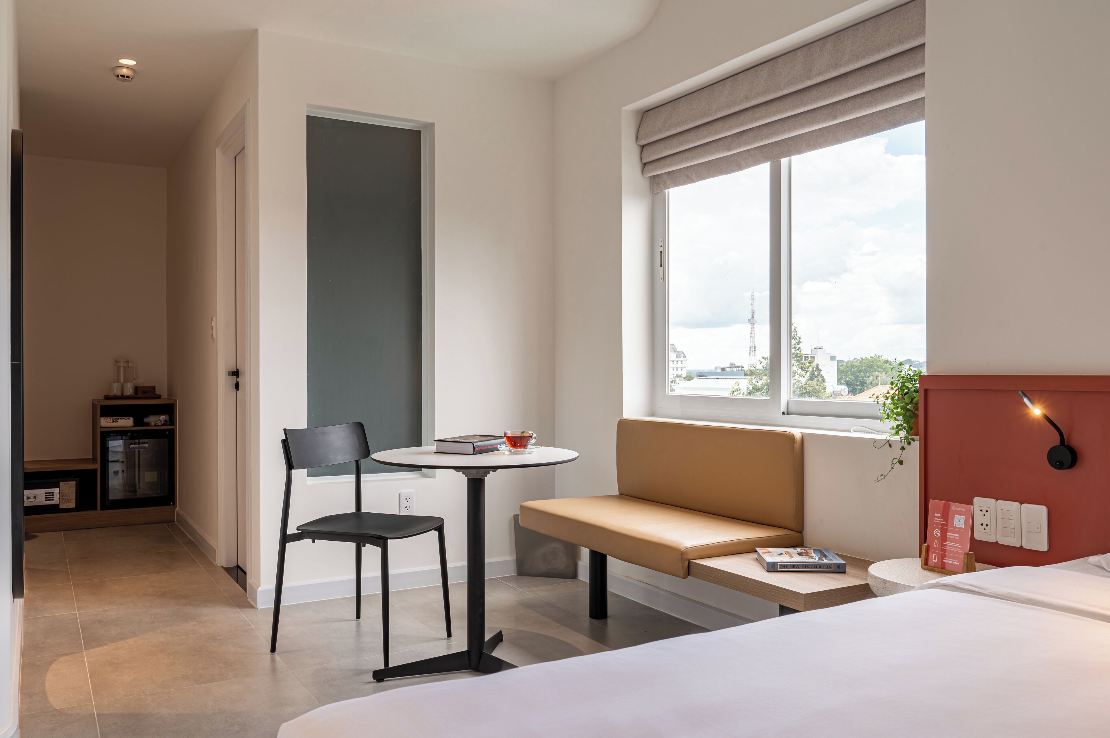 A modern hotel room with a bed, a small round table with a chair, a bench, a window with a view of the city, and a kitchenette area.