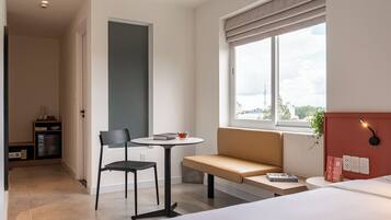 A modern hotel room with a bed, a small round table with a chair, a bench, a window with a view of the city, and a kitchenette area.