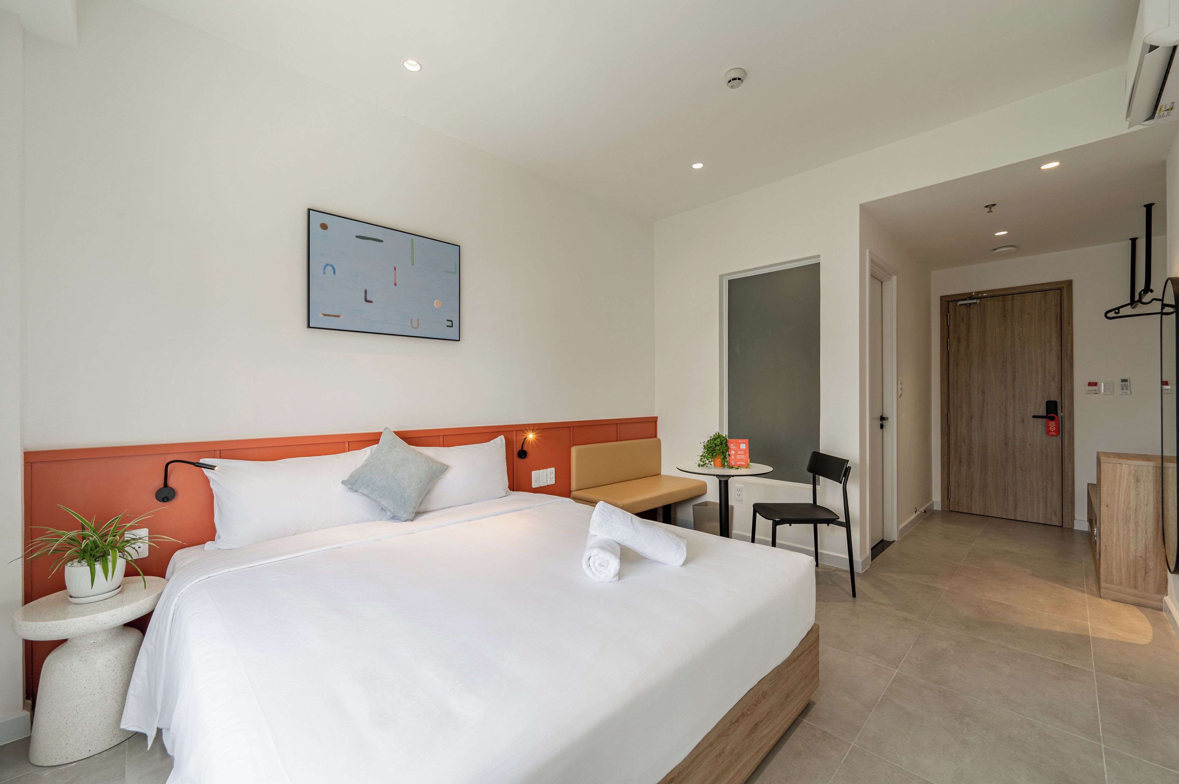 A modern hotel room with a large bed, a bedside table with a plant, a small table with a chair, and a door leading to a bathroom.