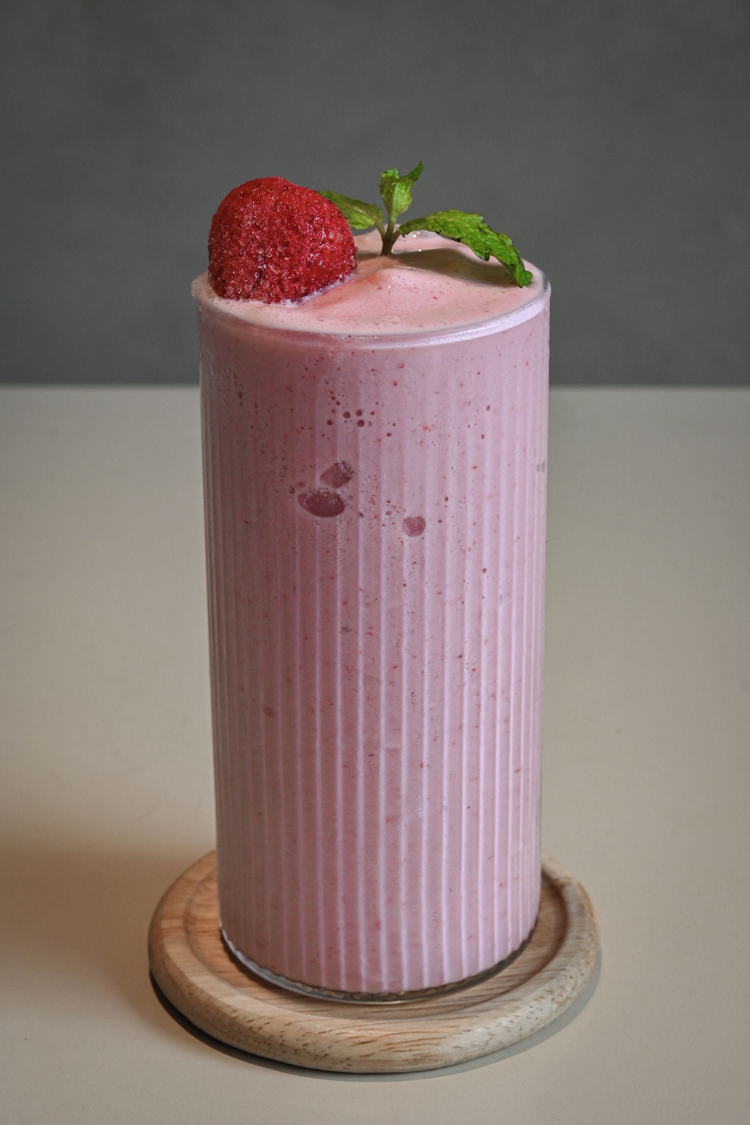 A tall, pink beverage in a glass with a strawberry garnish and mint leaf, placed on a wooden coaster.
