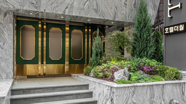 Ein Hotel Eingang mit einer Marmortreppe, die zu einer grünen und goldenen Tür führt, umgeben von einem angelegten Garten mit verschiedenen Pflanzen und einem Felsen.