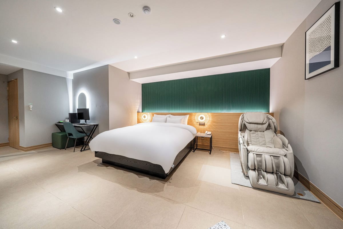 A modern hotel room with a large bed, a massage chair, a desk with a chair, and a green accent wall.